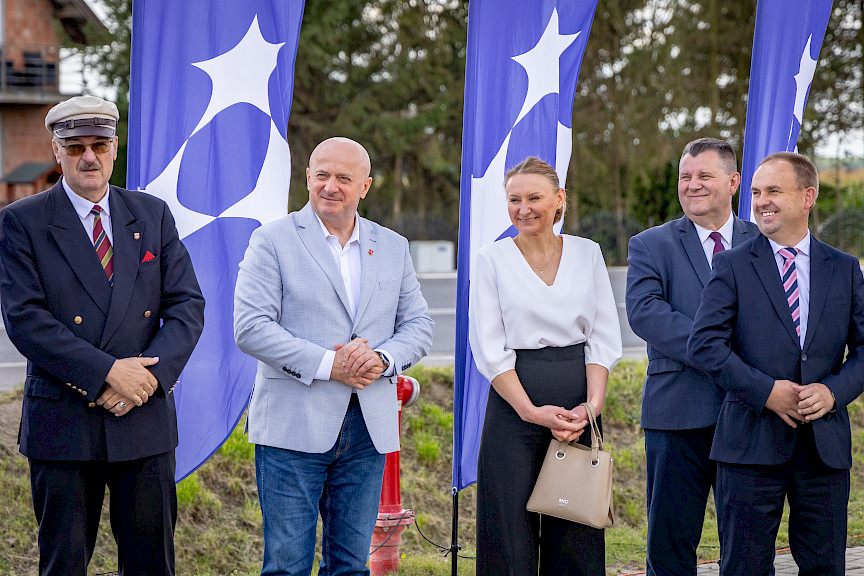 Burmistrz Urzędowa Paweł Dąbrowski stoi z Marszałkiem Jarosławem Stawiarskim oraz innymi osobami w galowych strojach. Uśmiechają się, a w tle widoczne są niebieskie flagi promujące fundusze europejskie dla lubelskiego.