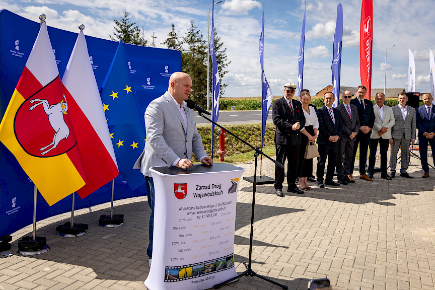 Marszałek Województwa Lubelskiego Jarosław Stawiarski wypowiada się do mikrofonu do uczestników wydarzenia. Obok stoją mężczyźni w garniturach oraz jedna kobieta. Za nimi stoją niebieskie flagi promujące fundusze europejskie dla lubelskiego.