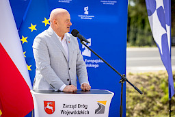 Marszałek Województwa Lubelskiego Jarosław Stawiarski wypowiada się do mikrofonu do uczestników wydarzenia. Za nimi stoją niebieskie flagi promujące fundusze europejskie dla lubelskiego.