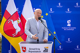 Marszałek Województwa lubelskiego Jarosław Stawiarski wypowiada się do mikrofonu do uczestników wydarzenia. Za nim stoi niebieska ścianka promująca fundusze europejskie dla lubelskiego..