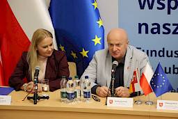 Marszałek Województwa Lubelskiego Jarosław Stawiarski wypowiada się do mikrofonu siedząc za stołem. Obok niego siedzi Dyrektor Departamentu Zarządzania Programami Regionalnymi Anna Brzyska.