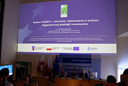 Pod sufitem na dużym ekranie wyświetlona jest informacja o systemie wprowadzonym w szpitalu. Pod ekranem widoczne są osoby siedzące za stołem oraz inni uczestnicy spotkania na sali.