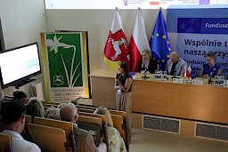 Ujęcie od góry sali na uczestników spotkania i kobietę wypowiadającą się do mikrofonu i wyświetlającą prezentację.