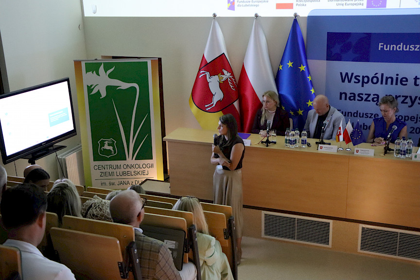 Ujęcie od góry sali na uczestników spotkania i kobietę wypowiadającą się do mikrofonu i wyświetlającą prezentację.