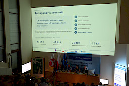 Pod sufitem wyświetlany jest duży obraz z prezentacją. Poniżej widoczni są siedzący na sali uczestnicy spotkania.