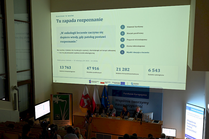 Pod sufitem wyświetlany jest duży obraz z prezentacją. Poniżej widoczni są siedzący na sali uczestnicy spotkania.