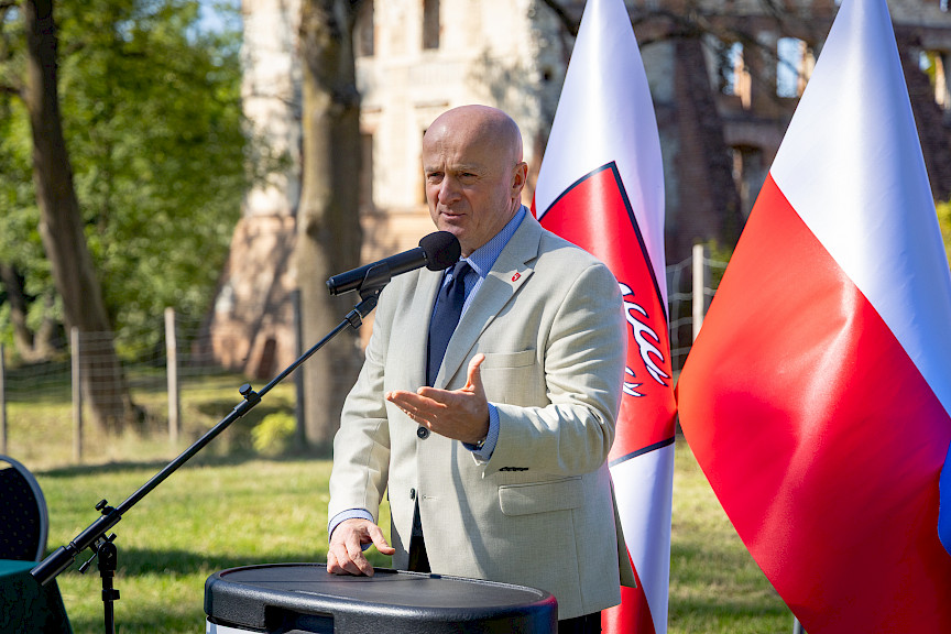 Marszałek Województwa Lubelskiego Jarosław Stawiarski przemawia do mikrofonu do uczestników wydarzenia. Za nim widoczne są flagi, Polski oraz Województwa Lubelskiego. Wydarzenie odbywa się na dworze.