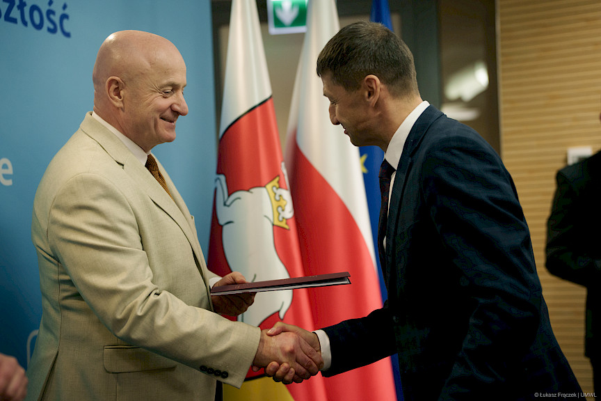 Marszałek Województwa Lubelskiego Jarosław Stawiarski podaje rękę w ramach gratulacji uśmiechając się beneficjentowi projektu. W drugiej ręce trzyma bordową teczkę z umową. Ujęcie z profilu.