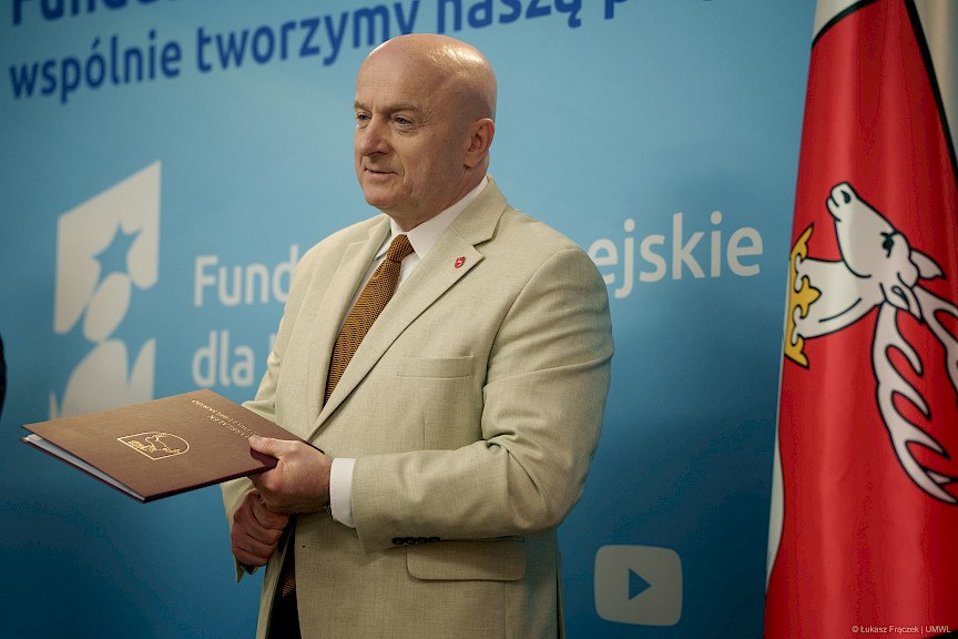 Marszałek Województwa Lubelskiego Jarosław Stawiarski stoi i oczekuje na podejście beneficjenta. W rękach trzyma bordową teczkę z umową.