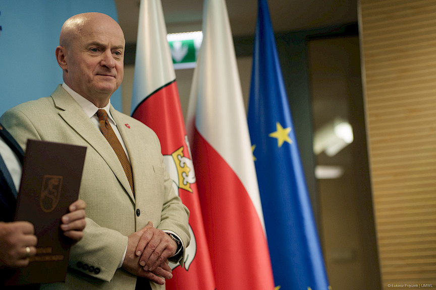Marszałek Województwa Lubelskiego Jarosław Stawiarski stoi i oczekuje na podejście beneficjenta. Za nim widoczne są flagi Unii Europejskiej, Polski i Województwa Lubelskiego.