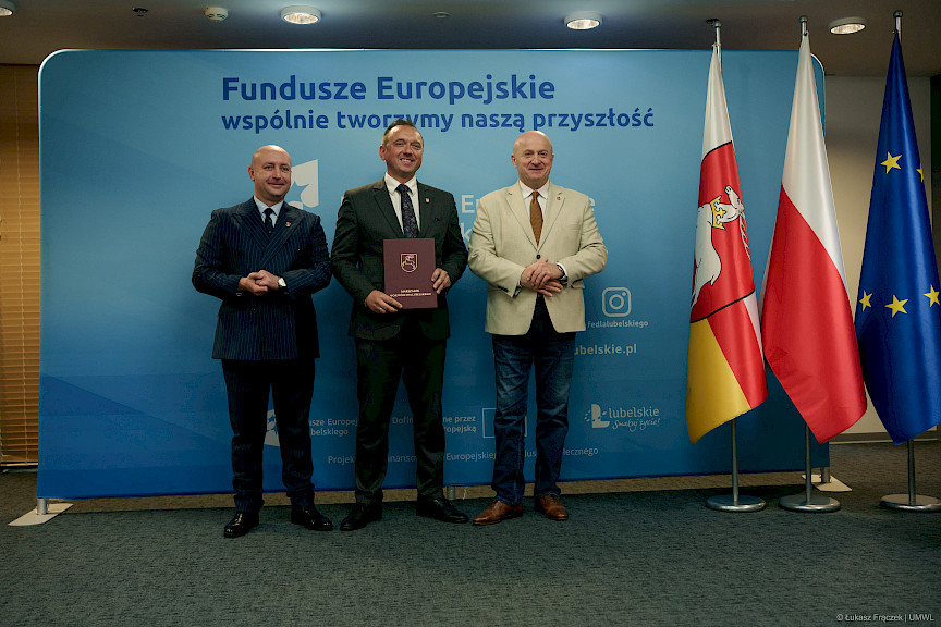 Marszałek Województwa Lubelskiego Jarosław Stawiarski oraz Wicemarszałek Piotr Breś pozują do zdjęcia. Pomiędzy nimi stoi beneficjent projektu, w rękach trzyma bordową teczkę z umową. Za nimi znajduje się niebieska ścianka promująca fundusze europejskie.