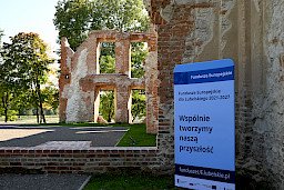 Ruiny zabytkowego budynku z czerwonej cegły i kamienia oraz niebieska tablica informacyjna 