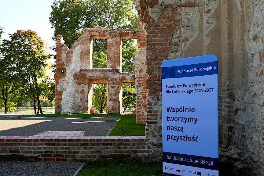 Ruiny zabytkowego budynku z czerwonej cegły i kamienia oraz niebieska tablica informacyjna 