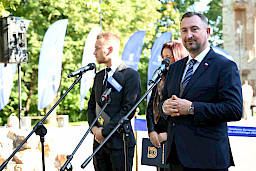 Wicemarszałek Województwa Lubelskiego Marek Wojciechowski przemawia o mikrofonu. Zdjęcie w plenerze, obok niego stoi kobieta oraz mężczyzna.