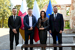 Wicemarszałek Województwa Lubelskiego Marek Wojciechowski stoi kobietą i mężczyznami pozując do zdjęcia na tle ruin pałacu oraz flag symbolizujących Polskę, Unię Europejską oraz Województwo Lubelskie.