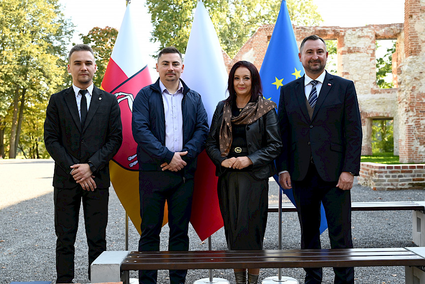 Wicemarszałek Województwa Lubelskiego Marek Wojciechowski stoi kobietą i mężczyznami pozując do zdjęcia na tle ruin pałacu oraz flag symbolizujących Polskę, Unię Europejską oraz Województwo Lubelskie.