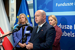 Wicemarszałek Województwa Lubelskiego Piotr Breś wypowiada się do mikrofonów. Za nim stoi Dyrektor Departamentu Zarządzania Programami Regionalnymi Anna Brzyska oraz druga kobieta. Ujęcie z profilu..