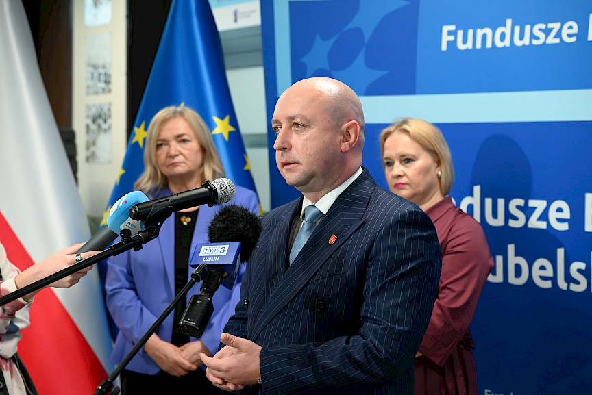 Wicemarszałek Województwa Lubelskiego Piotr Breś wypowiada się do mikrofonów. Za nim stoi Dyrektor Departamentu Zarządzania Programami Regionalnymi Anna Brzyska oraz druga kobieta. Ujęcie z profilu..