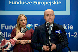 Wicemarszałek Województwa Lubelskiego Piotr Breś wypowiada się do mikrofonów. Za nim stoi Dyrektor Departamentu Zarządzania Programami Regionalnymi Anna Brzyska.