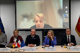 Prowadzący posiedzenie na czele z Wicemarszałkiem Bresiem siedzą za stołem i przysłuchują się wypowiedzi kobiety połączonej zdalnie, wyświetlonej na dużym ekranie,