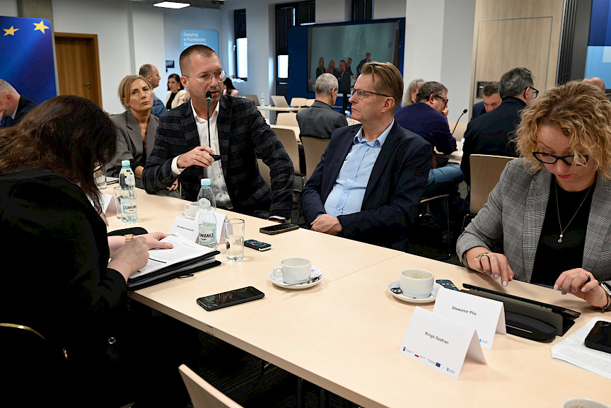 Duża liczba osób siedzących na sali przy stołach podczas posiedzenia Komitetu Monitorującego. Jeden z mężczyzn wypowiada się do mikrofonu z sali. 