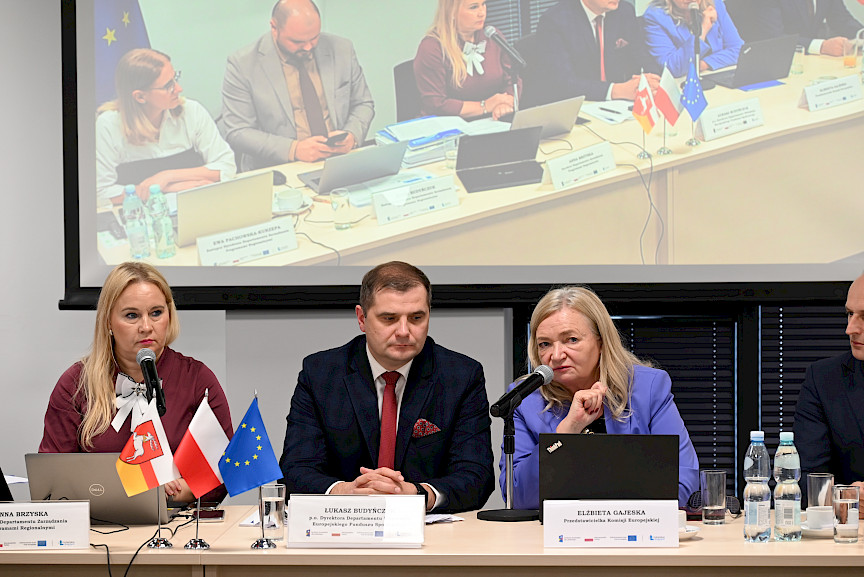 Przedstawicielka Komisji Europejskiej Elżbieta Gajeska wypowiada się do mikrofonu do uczestników spotkania. Koło niej siedzi Dyrektor Departamentu Wdrażania EFS Łukasz Budyńczuk oraz Dyrektor Anna Brzyska.