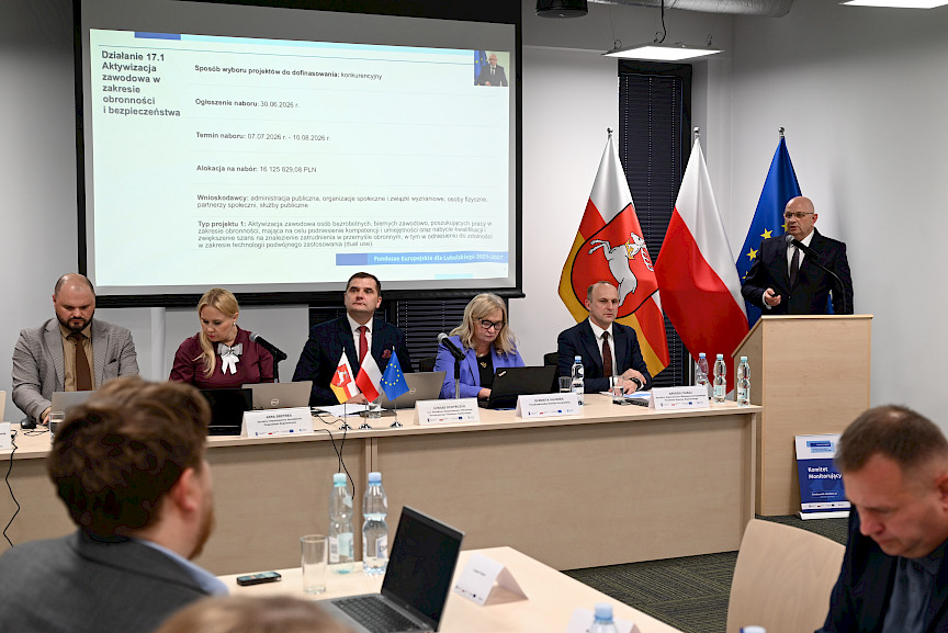 Mężczyzna wypowiada się przy ambonie do mikrofonu wyświetlając prezentację na dużym ekranie. Obok przy długim stole siedzą prowadzący spotkanie.