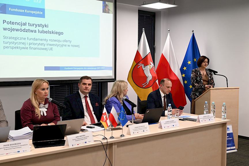 Kobieta wypowiada się przy ambonie do mikrofonu wyświetlając prezentację na dużym ekranie. Obok przy długim stole siedzą prowadzący spotkanie.