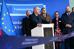 Wicemarszałek Województwa Lubelskiego Piotr Breś wypowiada się do mikrofonu. Stoi przy ambonie na dworze w płaszczu, za nim stoi kilka osób.