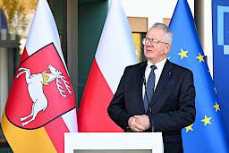 Mężczyzna w garniturze  wypowiada się przed budynkiem do mikrofonu do uczestników wydarzenia. Za nim znajdują się trzy flagi symbolizujące Polskę, Unię Europejską oraz Województwo Lubelskie.