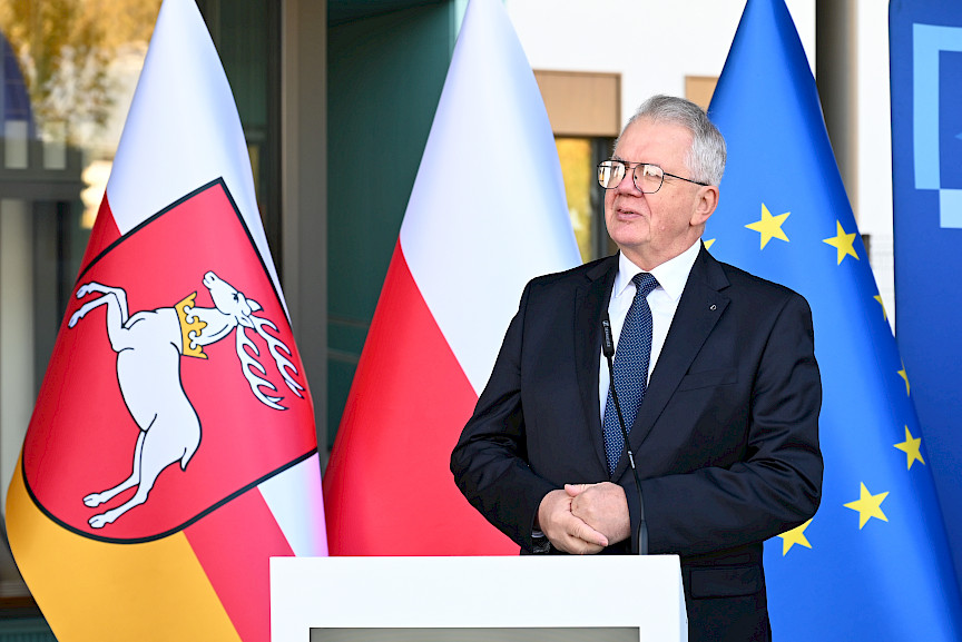 Mężczyzna w garniturze  wypowiada się przed budynkiem do mikrofonu do uczestników wydarzenia. Za nim znajdują się trzy flagi symbolizujące Polskę, Unię Europejską oraz Województwo Lubelskie.
