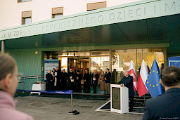 Wicemarszałek Województwa Lubelskiego Piotr Breś wypowiada się przed budynkiem do mikrofonu do uczestników wydarzenia. Pod dachem z logo szpitala stoją księża i kilka innych osób.