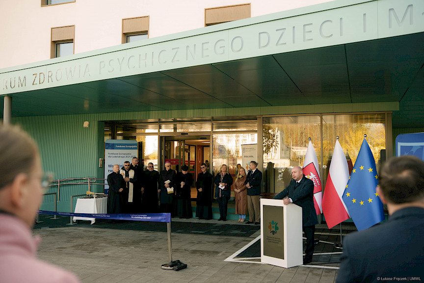 Wicemarszałek Województwa Lubelskiego Piotr Breś wypowiada się przed budynkiem do mikrofonu do uczestników wydarzenia. Pod dachem z logo szpitala stoją księża i kilka innych osób.
