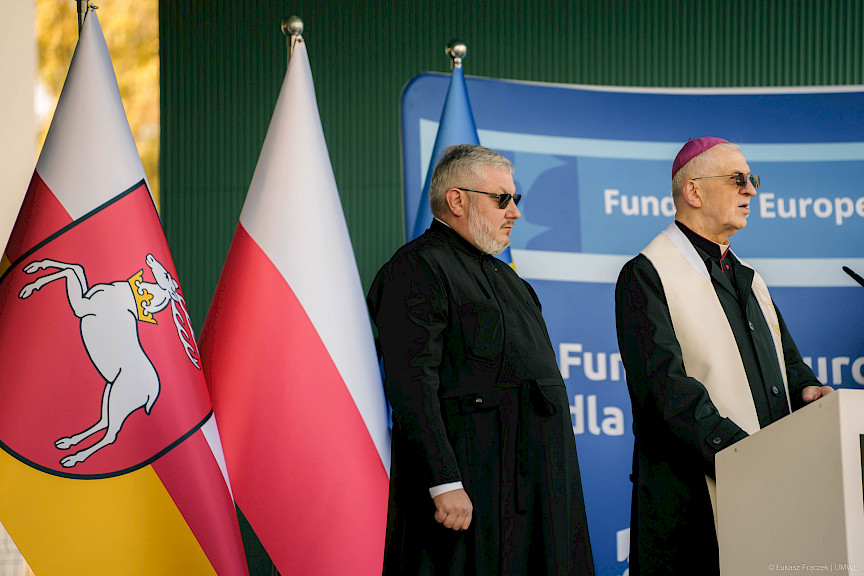 Ksiądz wypowiada się przed budynkiem do mikrofonu do uczestników spotkania. Obok niego stoi drugi ksiądz. Za nimi ustawione są flafi województwa lubelskiego oraz Polski.