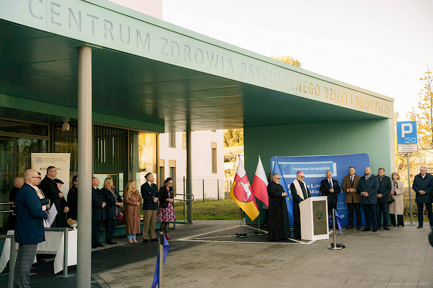 Grupa osób ustawionych pod dachem przed wejściem do szpitala podczas uroczystości.
