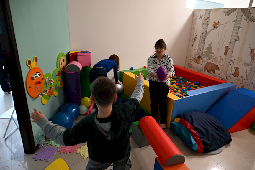 Sala terapeutyczna z basenem z kolorowymi kulkami, w której są dwie kobiety i chłopiec.